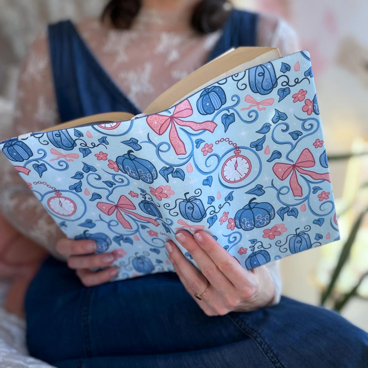 A women holding an open book with a light blue fabric dust jacket decorated with blue pumpkins, pinks bows and clocks .