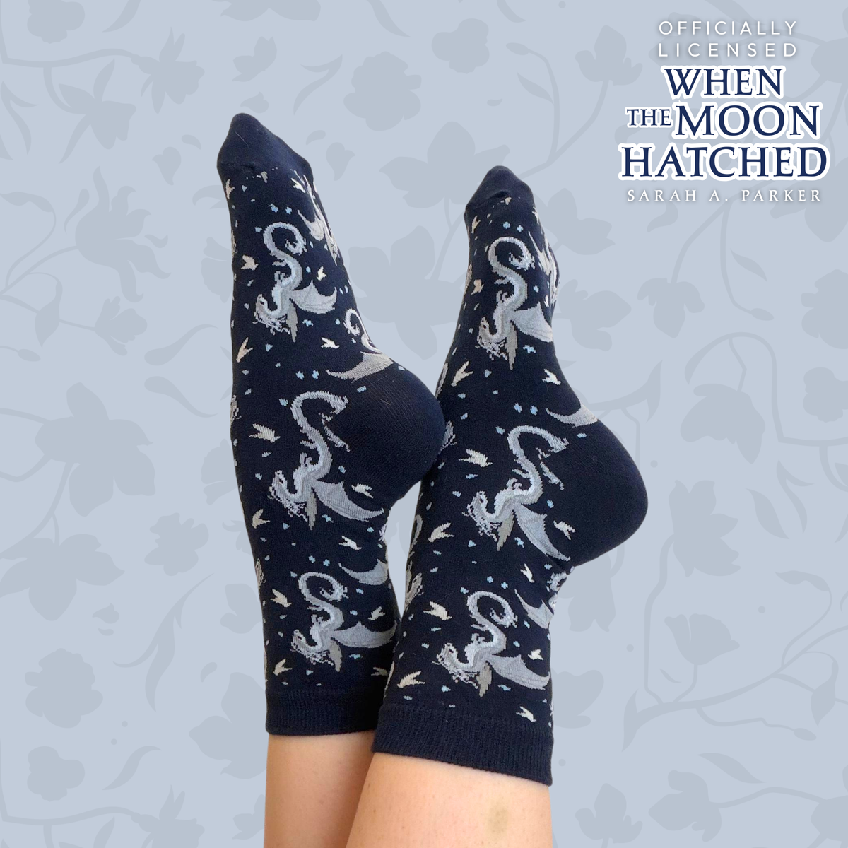 Navy blue socks featuring a pattern of white dragons against a light colored backdrop with a text overlay that reads "Officially licensed When the Moon Hatched by Sarah A. Parker"