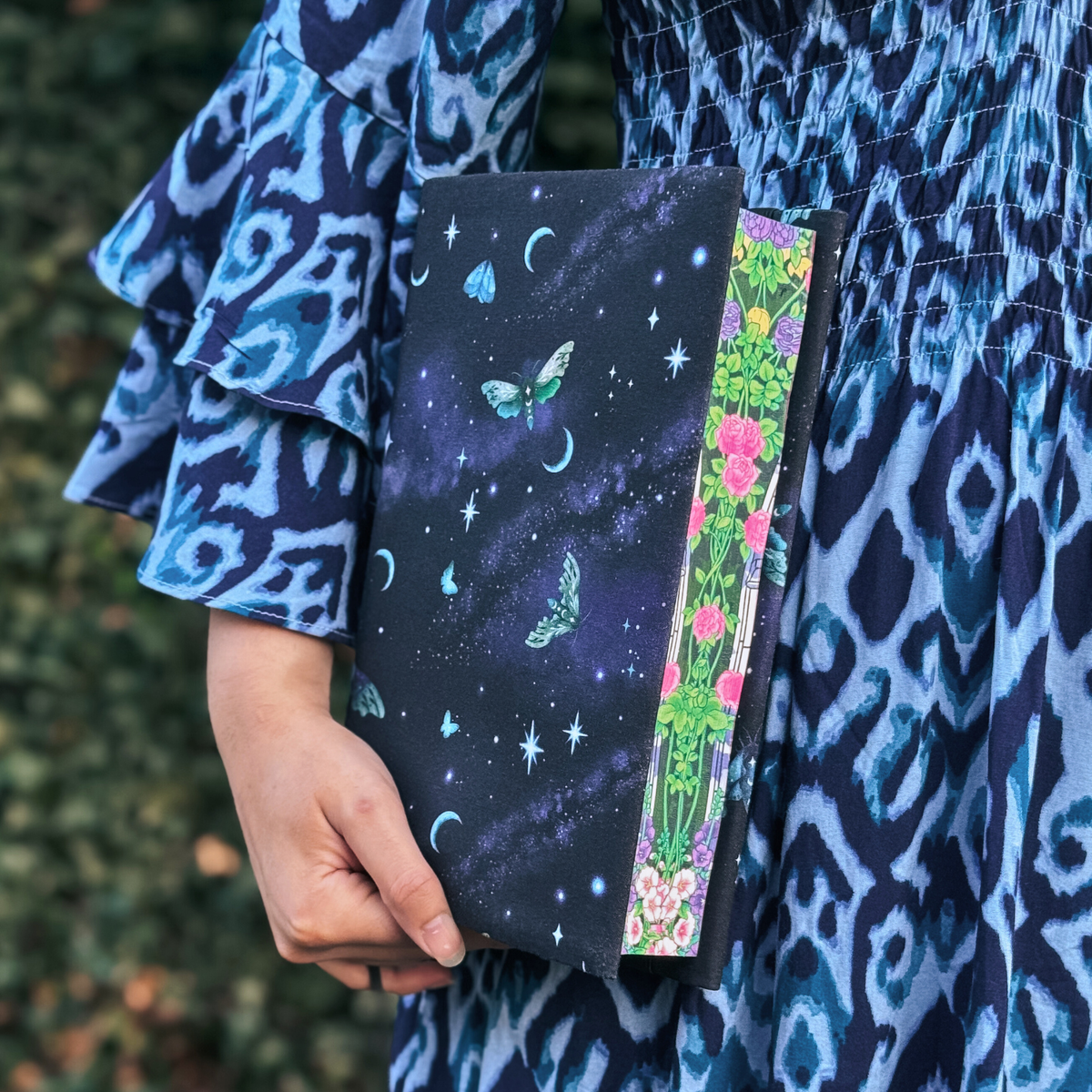A women holding a book with a dark blue fabric dust jacket decorated with light blue moths, stars, and moons against a blurred background.