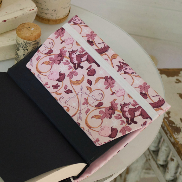 Close up of an open book with a light pink fabric dust jacket decorated with foxes, pink flowers light pink apples, bow and arrows, and gold swirls on top of a white table