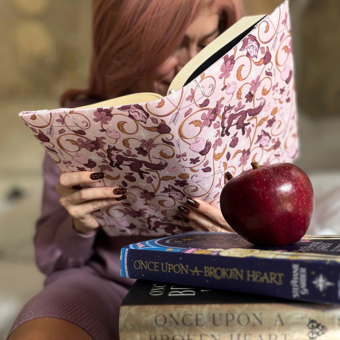 A women holding an open book with a light pink fabric dust jacket decorated with foxes, pink flowers light pink apples, bow and arrows, and gold swirls sitting in front of a stack other books with a red apple on top