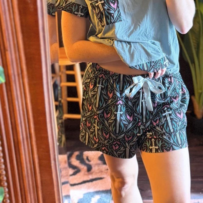 A women wearing pajama shorts with a pattern of swords, swirls, and leaves in colors of teal, light blue, and pink with a matching light blue t-shirt