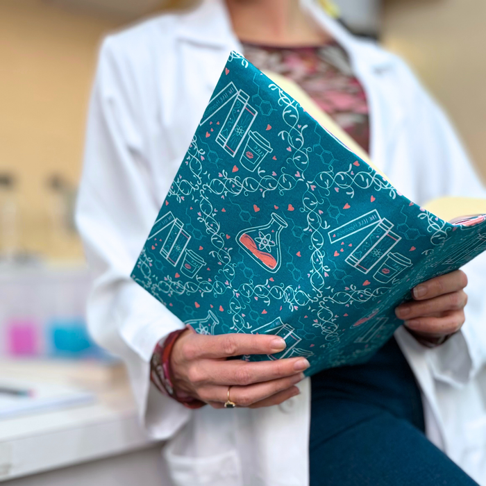 A women in a white lab coat holding an open book with a teal fabric dust jacket decorated with white scientific illustrations, including beakers, test tubes, DNA helixes, and pink hearts against a softly blurred laboratory background.