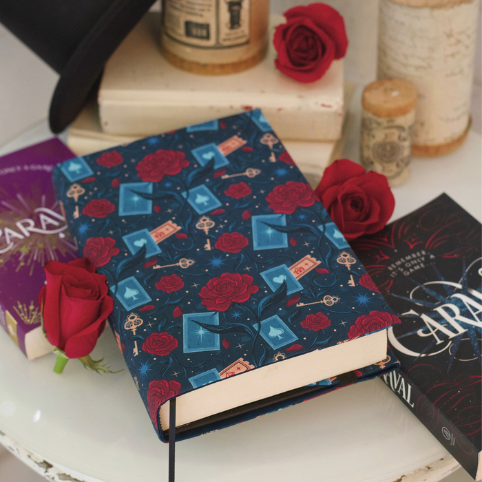A book with a dark blue fabric dust jacket decorated with red roses, blue playing cards, and golden keys and tickets on a white table with two copies of the book Caraval on it