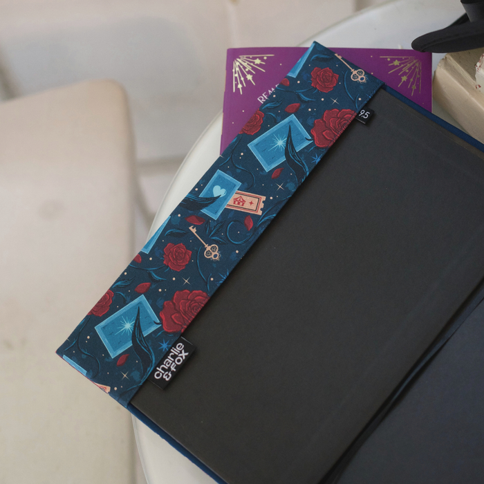 A close up of an open book with a dark blue fabric dust jacket decorated with red roses, blue playing cards, and golden keys against a blurred white room