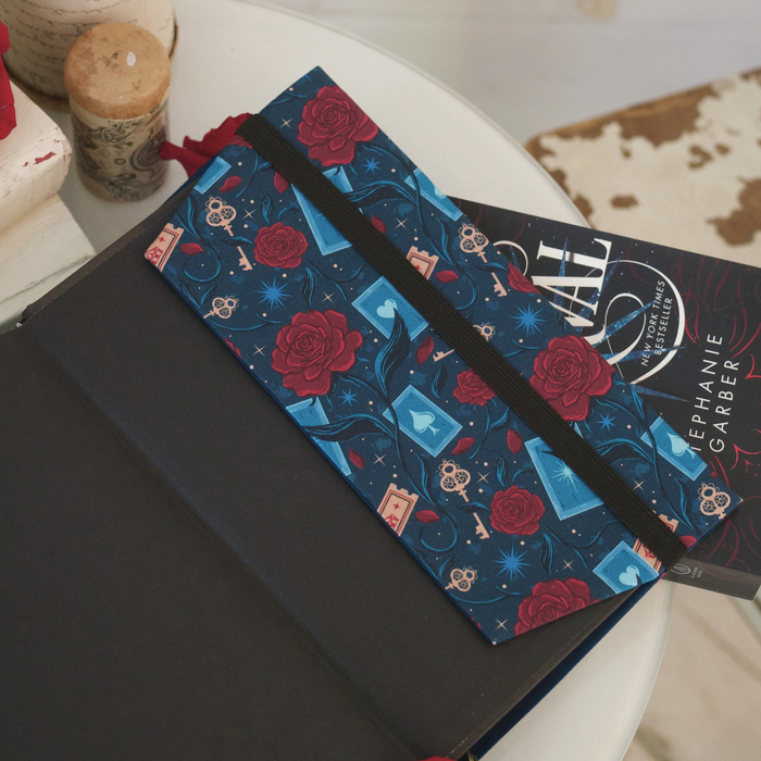 A close up of an open book with a dark blue fabric dust jacket decorated with red roses, blue playing cards, and golden keys sitting on top of another book on a white table