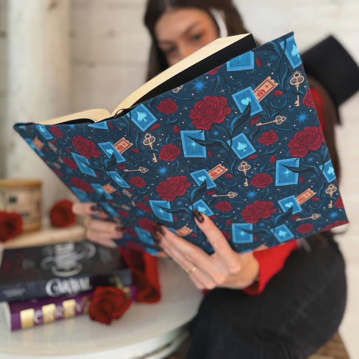 A women holding an open book with a dark blue fabric dust jacket decorated with red roses, blue playing cards, and golden keys sitting at a table with two copies of the book Caraval on it