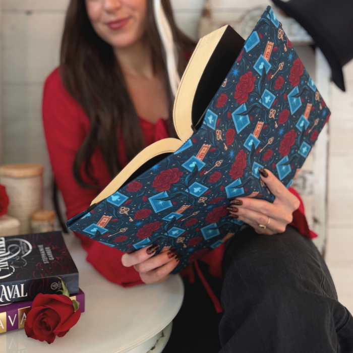 A women holding an open book with a dark blue fabric dust jacket decorated with red roses, blue playing cards, and golden keys sitting at a white table with other books on it.
