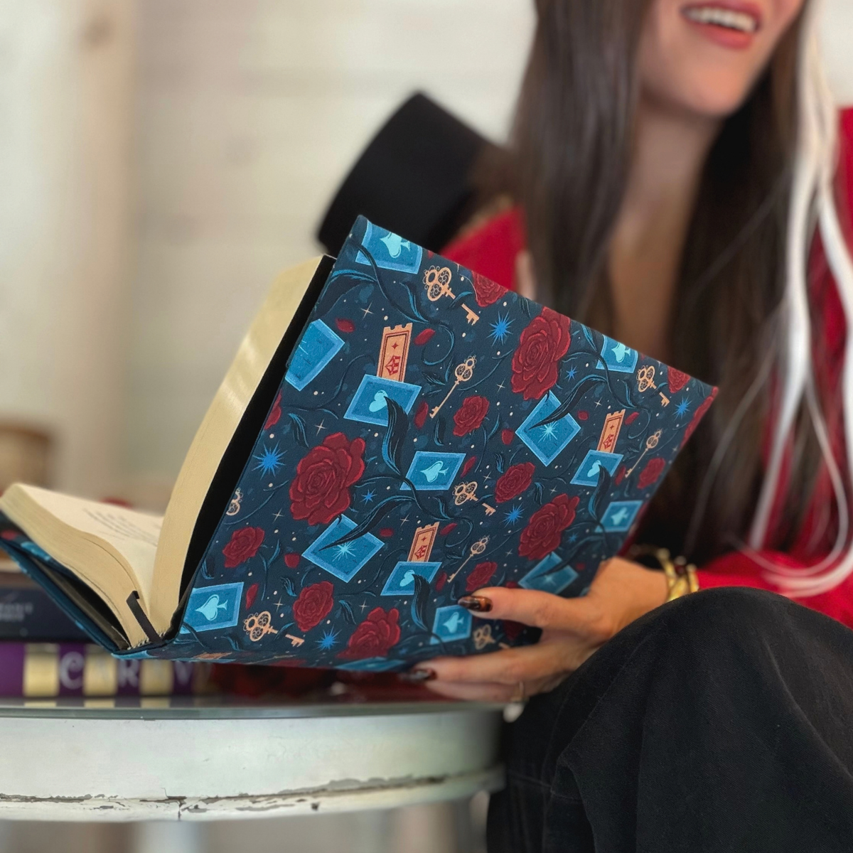 A women holding an open book with a dark blue fabric dust jacket decorated with red roses, blue playing cards, and golden keys sitting at a white table with other books on it.
