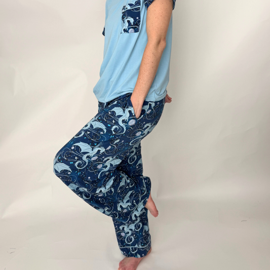A women wearing a pajama set with a light blue t-shirt and blue pants with a pattern of light blue dragons, moons, and constellations standing in front of a white background  