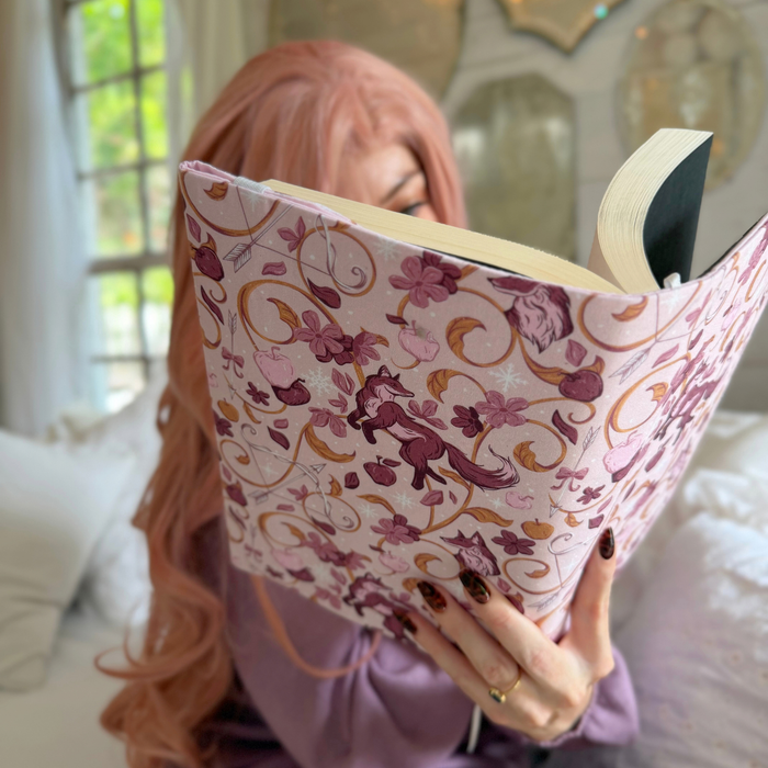 A women holding an open book with a light pink fabric dust jacket decorated with foxes, pink flowers light pink apples, bow and arrows, and gold swirls sitting on a white bed