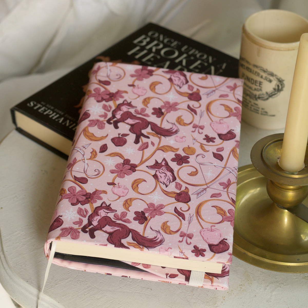 A book with a light pink fabric dust jacket decorated with foxes, pink flowers light pink apples, bow and arrows, and gold swirls on a white table with another book