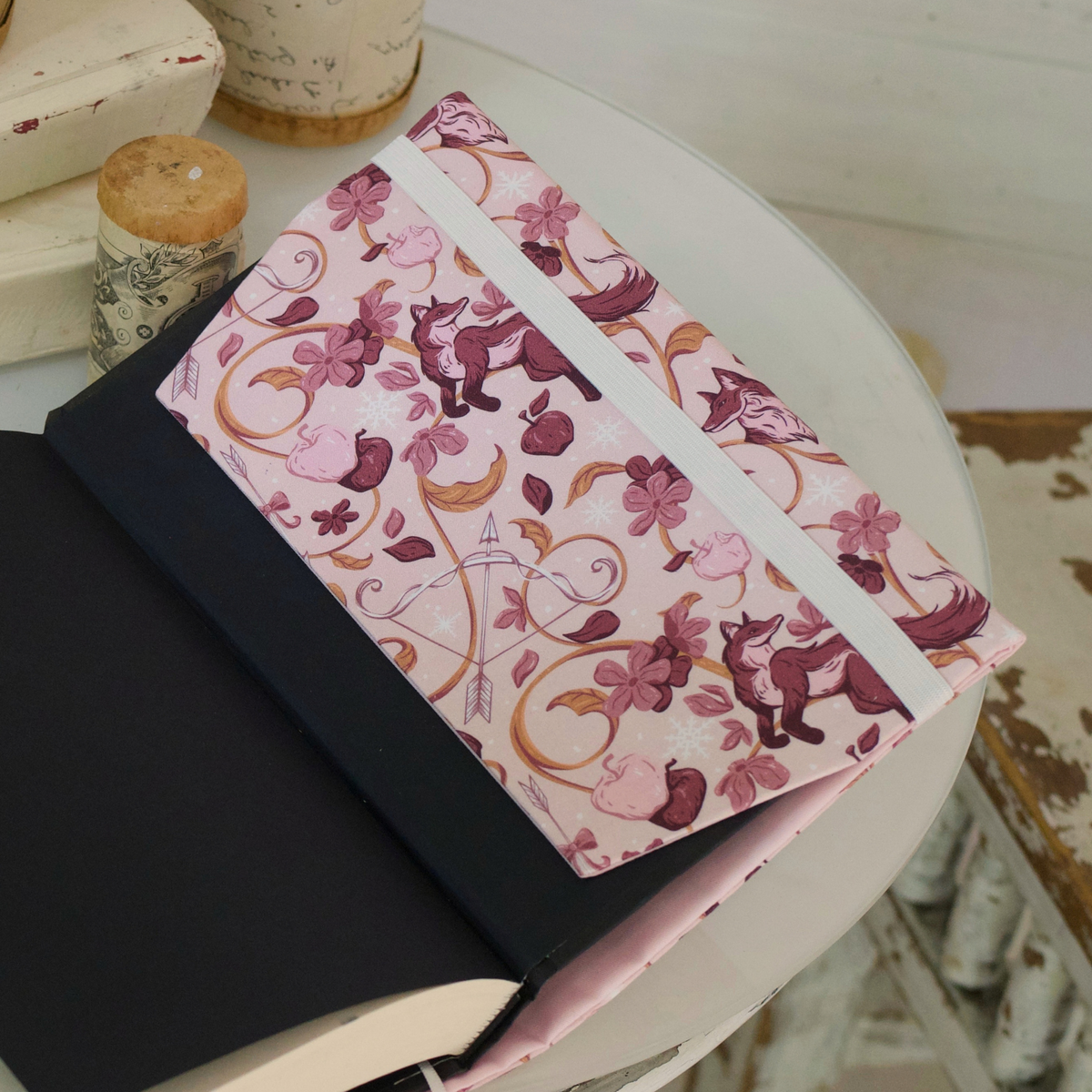 Close up of an open book with a light pink fabric dust jacket decorated with foxes, pink flowers light pink apples, bow and arrows, and gold swirls on top of a white table
