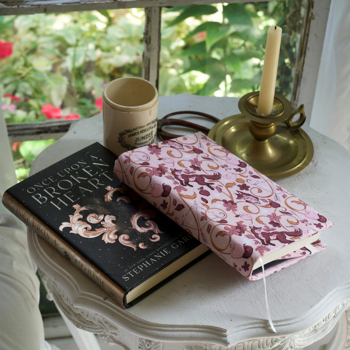 A book with a light pink fabric dust jacket decorated with foxes, pink flowers light pink apples, bow and arrows, and gold swirls on a white table with another book titled Once Upon A Broken Heart