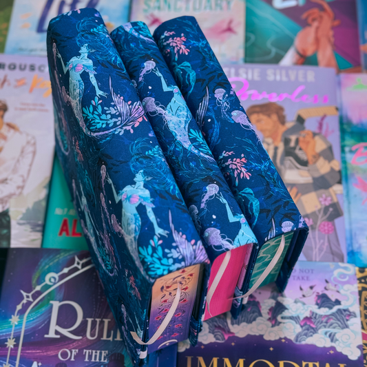 A stack of books with  a blue fabric dust jacket decorated with mermaids, light purple jellyfish, and pink aquatic plants sitting on top of a table covered in other books. 