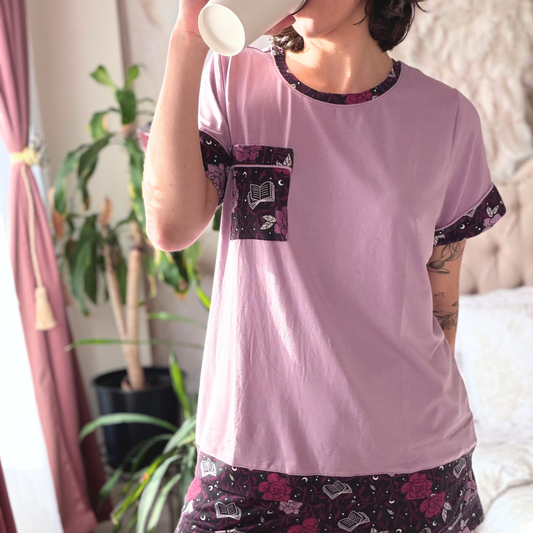 A women wearing a pajama set of a muted purple t-shirt and shorts with a floral pattern of pink and muted purple roses drinking a coffee 