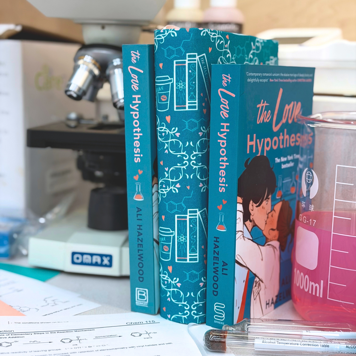 A book with a teal fabric dust jacket decorated with white scientific illustrations, including beakers, test tubes, DNA helixes, and pink hearts in between two coped of the book The Love Hypothesis  on a science bench with beakers and a microscope on it. 
