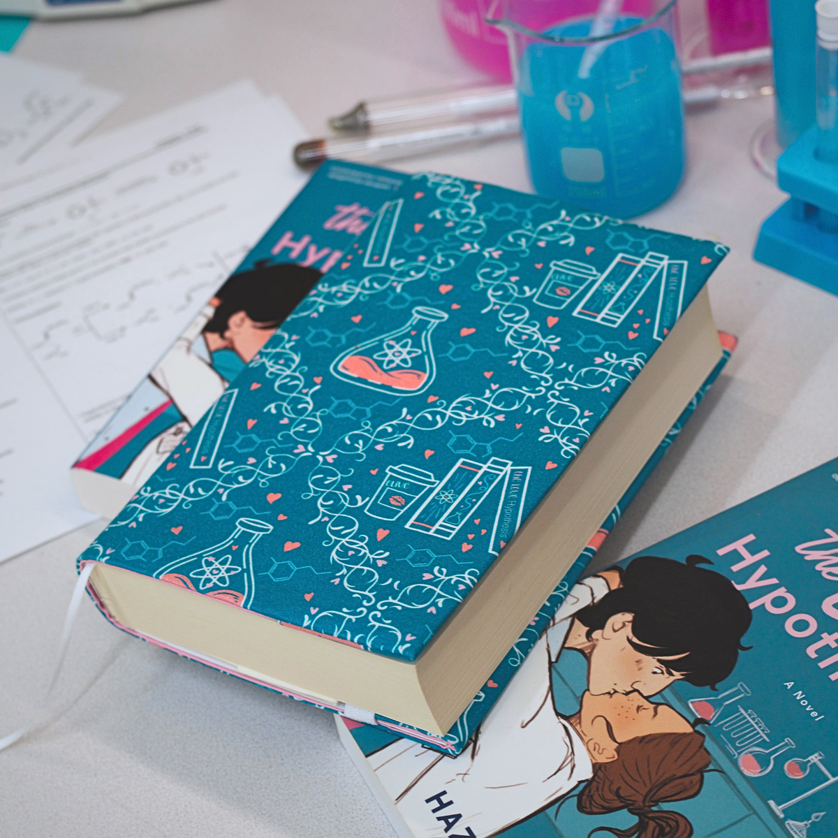 A book with a teal fabric dust jacket decorated with white scientific illustrations, including beakers, test tubes, DNA helixes and pink hearts, on top of two copies of the book The Love Hypothesis with blurred test beaker in the background