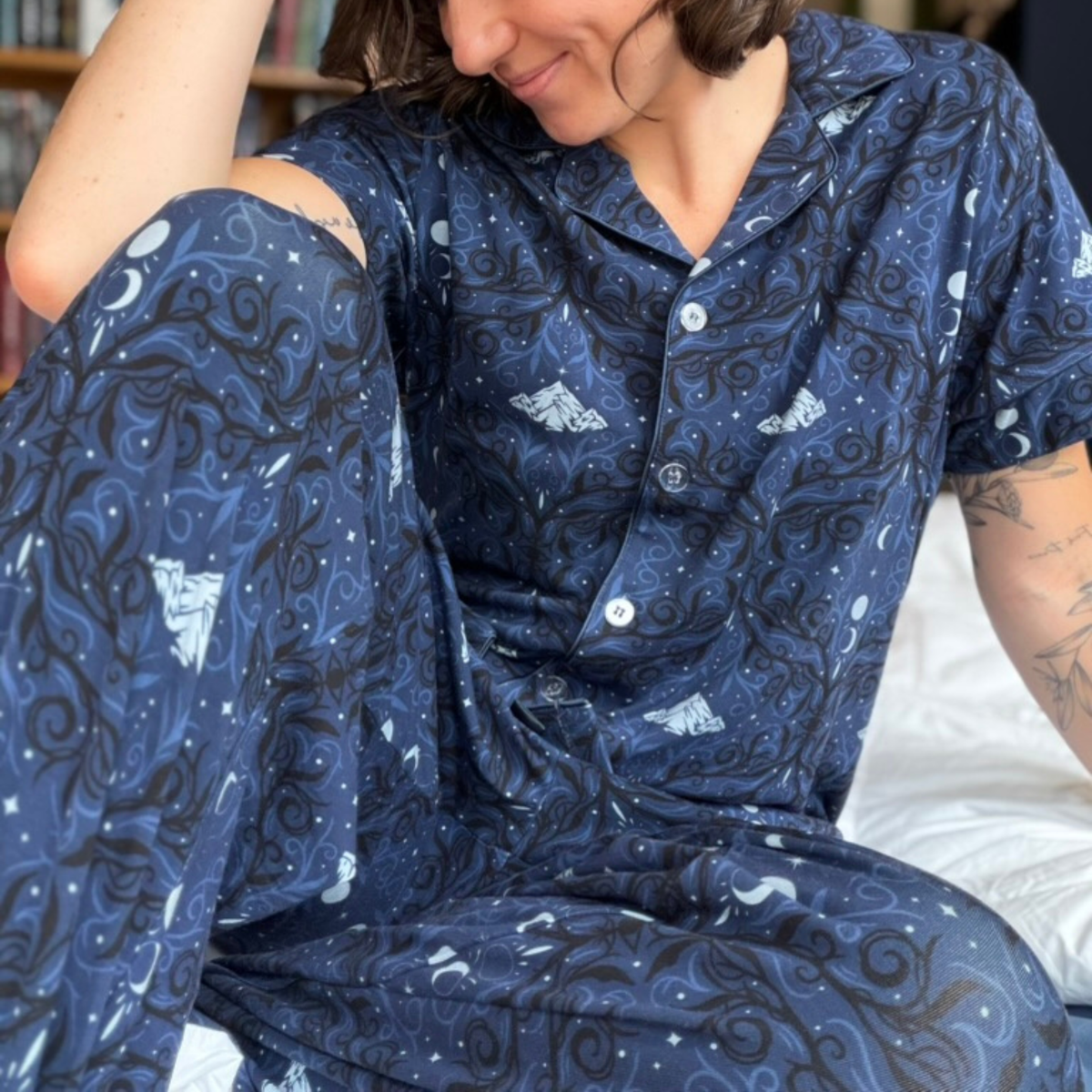 A women wearing a navy blue pajama set of a short sleeve button-up shirt and pants with a pattern of white mountains and moons and light blue and black swirls sitting on a white blanket. 