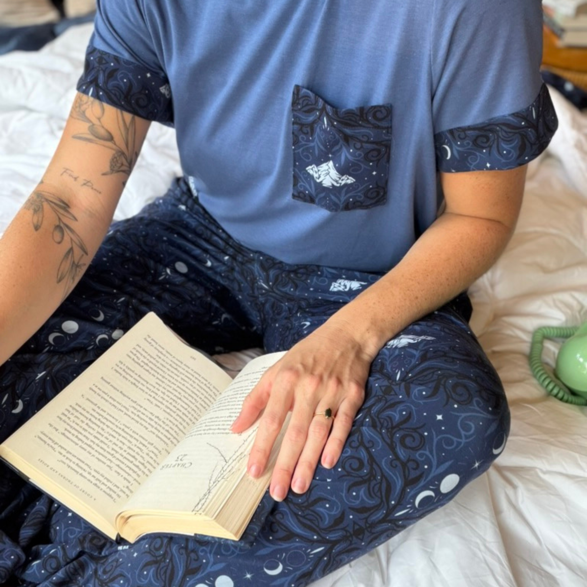 A women wearing a pajama set of a blue t-shirt an navy blue pants with a pattern on white mountains and moons and light blue and black swirls sitting on a white blanket with a book in her lap