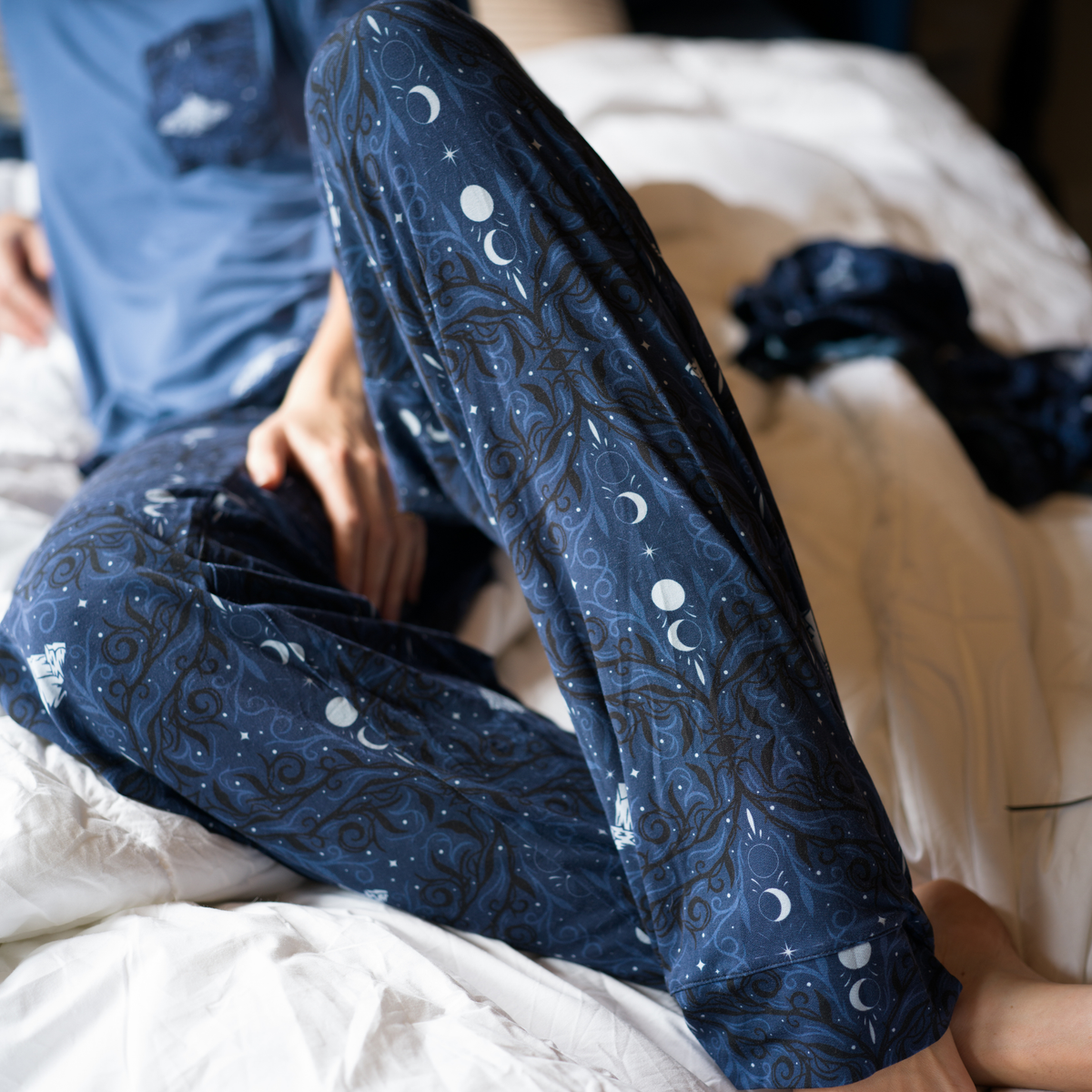 A women reclining on a bed wearing navy blue pajama pants with white moons and mountains and black and light blue swirls 